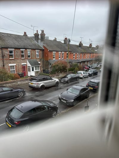 The image displays a residential street scene in Dorchester, United Kingdom, viewed from an elevated perspective, likely a window, with a vertical metallic obstruction partially obscuring the right frame. The environment is overcast and appears wet from rain.
The street, identified as a residential road, features terraced houses constructed primarily of red brick with light-colored window frames, dark grey tiled roofs, and prominent chimneys and television aerials. Several houses have small front gardens with low brick walls and sparse autumn foliage. Black refuse bins are visible outside some properties. Overhead power or communication lines crisscross the street.
A utility pole stands near the center of the street, positioned between the two rows of houses. An individual, wearing dark clothing and possibly safety gear, is observed on the utility pole, appearing to work on the overhead lines.
Numerous vehicles are parked on both sides of the street. Prominently visible in the mid-ground are a silver Range Rover Sport (UK registration Y16 KNS), a black BMW sedan (UK registration RJ66 BFV), a dark Citroën DS3 hatchback (UK registration CE67 FPG), and a dark Mercedes-Benz coupe (UK registration ME65 XUC). In the foreground, a dark hatchback (likely a SEAT Ibiza, UK registration SJ17 N A) is parked on the left. Further down the street, a silver Volkswagen hatchback, a black Mini Cooper, a white van, and other cars are visible. The road surface is wet, reflecting the grey sky. White line markings delineate parking areas. No other people are clearly discernible.