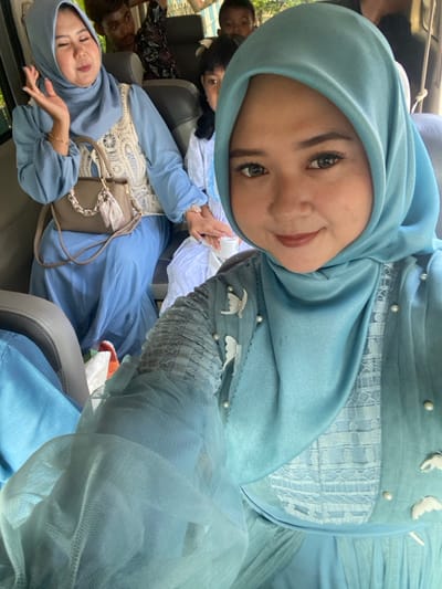 The image depicts five individuals inside a vehicle in Ketegan, Indonesia. In the immediate foreground, a woman in a light blue hijab and a matching dress adorned with white butterfly-shaped embellishments and pearls is taking a selfie, looking directly at the camera with a subtle smile. Her right arm, showcasing intricate sleeve detailing with sheer fabric and gathered sections, is visible on the lower left. Directly behind her, in the mid-ground, another woman in a light blue hijab and dress is seated. She wears a cream-colored, open-knit vest over her dress and holds a light brown handbag with a silver chain strap and a decorative tassel on her lap. Her right hand is raised, palm open, in a gesturing motion. Behind these two women, partially obscured, are three other individuals: a young child with dark hair positioned between the two foreground women, looking towards the camera; a person with dark, short hair visible behind the mid-ground woman; and another child's face partially visible in the upper right background. The interior of the vehicle, characterized by visible seatbacks and muted tones, suggests travel or transport.