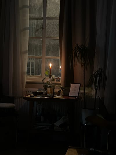 A dimly lit room features a window covered with sheer curtains, through which an exterior scene is visible. A table sits in front of the window, adorned with a burning candle and what appears to be a botanical arrangement within a glass container. A framed picture or document is positioned on the table. To the right, a chair is partially visible, merging into the shadowed background. A potted plant stands atop the table, with its silhouette extending towards the ceiling.
