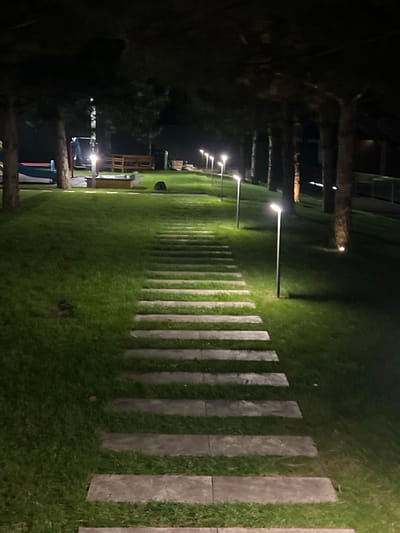 The image shows a night-time view of a well-maintained, grassy yard with a stone pathway leading into the distance. The pathway is made of evenly spaced, rectangular stone slabs laid horizontally across the lawn. Along the pathway, modern, thin lampposts with upward-facing lights illuminate the scene, casting a soft glow on the grass and trees. The trees, which appear to be pines due to their conical shape and dark foliage, are positioned on either side of the yard, creating a tunnel-like effect. In the background, there are glimpses of playground equipment, including a slide and what might be a sandbox, as well as wooden benches and a fence. The overall impression is one of a tranquil, illuminated outdoor space, likely a park or a residential backyard, in the city of Vyshen'ky, Ukraine. There are no people visible in the image.