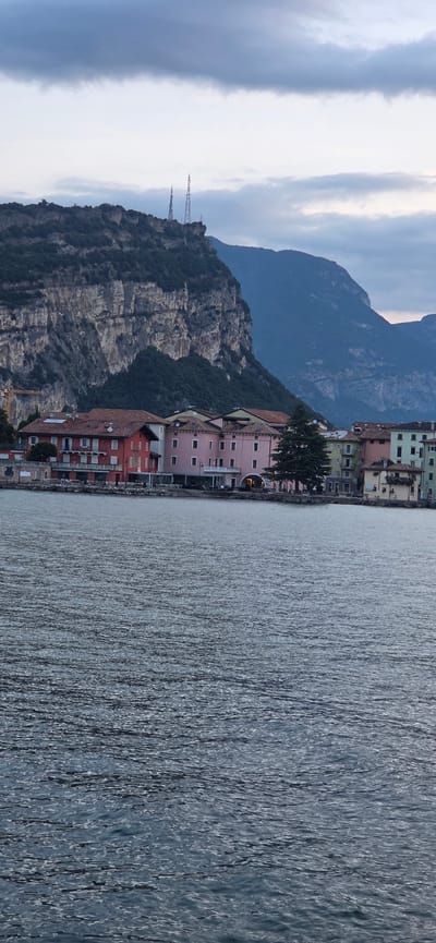 The image captures a waterfront scene in Torbole, Italy, on an overcast day. In the foreground, the rippling surface of a body of water occupies the lower half of the frame. Beyond the water, a row of buildings lines the shore. The architecture is varied, with several multi-story structures featuring pitched roofs. Some buildings are painted in warm tones like red and pink, while others are lighter in color, such as pale yellow and green. A large, dark green evergreen tree stands prominently among the buildings.
Behind the town, a massive, steep, rocky cliff face dominates the background, its surface textured and weathered. The upper edges of the cliff are covered with dark green vegetation. Two tall, lattice-like radio towers are visible on the crest of the cliff, silhouetted against the cloudy sky. Further in the distance, the soft outlines of more mountains recede into the horizon. The sky is a muted grey with patches of lighter clouds. No people are visible in the scene. The overall impression is one of a tranquil, natural landscape meeting a settled, historical town.