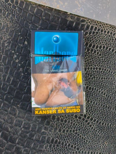 The image shows a top-down view of a pack of Marlboro Ice Blast Mega cigarettes placed on a textured, dark surface that appears to be a table or countertop with a snakeskin-like pattern. The cigarette pack is predominantly blue and silver, featuring the Marlboro logo and a graphic of a circular blue lens with a white dot in the center.

The front of the pack displays a shocking health warning graphic, as mandated by law in the Philippines. This graphic depicts a close-up view of a breast with an open wound, likely representing the detrimental effects of smoking. Below this graphic, in yellow text with a black outline, are the words "ANG PANINIGARILYO AY SANHI NG KANSER SA SUSO," which translates to "SMOKING CAUSES BREAST CANCER" in English.

The overall scene is static, with no specific activity or event taking place beyond the presentation of the product and its accompanying warning. The lighting appears to be natural, suggesting it was taken during the daytime, and there are no discernible weather cues. The presence of the health warning in Tagalog strongly indicates the location context is the Philippines, specifically Cavite City. The mood conveyed by the health warning graphic is somber and cautionary, aiming to deter smoking.