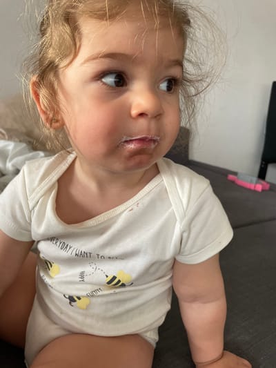 A young child, approximately one to two years old, with light brown, wavy hair and dark eyes, is depicted from the upper torso. The child wears a white, short-sleeved onesie featuring the text "EVERYDAY I WANT TO BEE..." and yellow bumblebee illustrations. White residue, consistent with a dairy product, is visible on the child's upper lip and chin. Minor food stains are present on the onesie. The child is looking towards the right of the frame with a focused gaze. The background shows a light-colored wall on the left and a dark grey upholstered surface, likely a sofa, extending to the right and beneath the child. A pink and grey plastic object rests on the sofa behind the child's right shoulder. This scene is located within a residential interior in Senden, Germany.