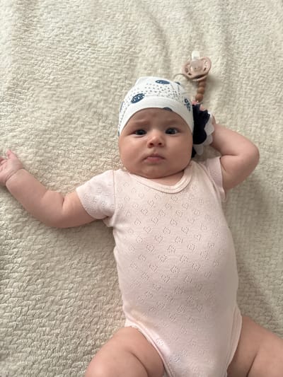 A baby lies on a cream-colored, textured blanket. The baby wears a light pink, short-sleeved onesie with a subtle diamond pattern and a white cap adorned with blue fish and silver accents. A pacifier clip with a wooden bead and a light pink pacifier is attached to the hat. The baby's left arm is extended, and the right arm is bent, with a hand near the head. The background is the blanket, and there is no indication of the city of Serebryakovka, Russia.