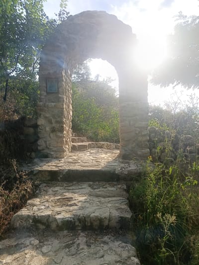 This image depicts an ancient stone archway, a pathway paved with rough-hewn stones leading up to it, and a series of stone steps disappearing beyond. The archway is constructed from large, irregular stones and frames a view of lush green foliage and trees bathed in bright sunlight. A small, framed picture, possibly an icon, is mounted on the left pillar of the archway. The scene is outdoors, set within a natural, overgrown environment, suggesting a historical or spiritual site. The sun is high in the sky, casting strong light and creating lens flares, indicating it is likely midday. The bright sunlight and dappled shadows suggest fair weather. The stone construction and the overall ambiance hint at a location with a long history, consistent with Melnik, Bulgaria, known for its ancient ruins and churches. No visible text is present in the image.