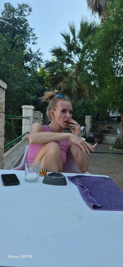 A woman with blonde hair pulled up in a bun, wearing sunglasses on her head and a pink tank top, is seated outdoors on a white chair. She is smoking a cigarette, with her left hand resting on her chin and her right hand holding the cigarette. In front of her, on a white table, is a smartphone, a glass with a white substance, a gray ashtray with cigarette butts, and a purple towel. The background features greenery, palm trees, and a clear blue sky. There are stone pillars and a railing in the mid-ground. The scene appears to be in a residential or outdoor dining area, possibly Almáchar, Spain, given the vegetation and architectural elements.