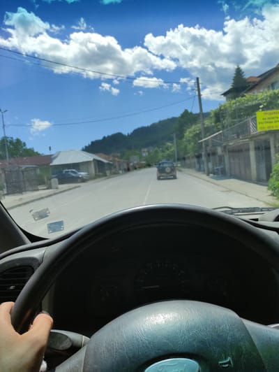 The image is a point-of-view shot from the driver's seat of a car, looking through the windshield. The scene depicts a street in Rudozem, Bulgaria, on a sunny day with scattered clouds. The road is paved and appears to be a main thoroughfare in a town or village. On either side of the road are buildings, some with tiled roofs and others with more modern structures. Green trees and vegetation are visible, suggesting a hilly or mountainous landscape in the background. A red SUV is driving ahead of the car. The steering wheel of the car is in the foreground, and a person's hand is visible on the left side of the wheel. A yellow sign with Bulgarian text is attached to a building on the right side of the road. The overall impression is of a scenic drive through a European town.