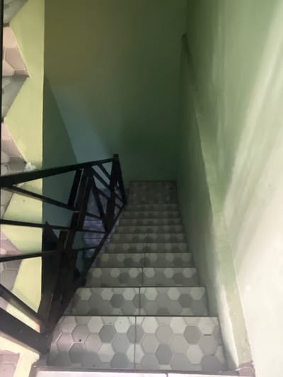 The image depicts a downward view of an indoor staircase with light green walls. The stairs are tiled with hexagonal patterns in shades of grey and white. A black metal railing lines the left side of the staircase. The overall environment suggests an interior space, possibly within a building in Pasir Gadung, Indonesia, though no specific landmarks or context confirm this location. The scene is static and there are no people or animals present. The lighting appears to be artificial and ambient, casting subtle shadows.