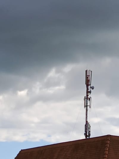 The image shows a telecommunications tower situated atop a red-tiled roof, with a backdrop of a cloudy sky. The tower, constructed from metal, is equipped with multiple antennas and circular dishes, suggesting it's a significant communication hub. The sky is overcast with dramatic, dark clouds dominating the upper portion of the frame, hinting at approaching rain or a stormy atmosphere. Below the dense clouds, lighter, diffused clouds are visible, and patches of bright sky can be seen. The red-tiled roof extends across the bottom of the image, providing a grounding element to the scene. There are no people visible in the image. The lighting suggests it could be late afternoon or early evening, or simply a day with heavy cloud cover. The overall mood is somewhat somber due to the weather conditions. The presence of the tower and the roof structure indicates an urban or semi-urban environment, likely residential or commercial. No text is visible in the image.