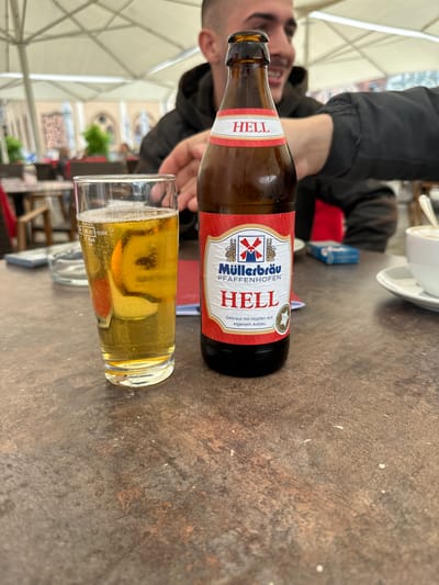 An outdoor table at a cafe in Pfaffenhofen an der Ilm, Germany, holds a glass of beer with a slice of citrus fruit and a bottle of Müllerbräu Hell beer. A person's arm is visible reaching towards the bottle. In the background, a blurred image shows an outdoor seating area with patrons and umbrellas. The table surface is a textured, dark brown material.