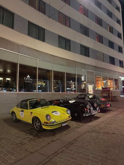 A nighttime scene depicts three vintage automobiles parked on a stone-paved surface in front of the Crystal Hotel in Sankt Moritz, Switzerland. The building's ground floor features large glass windows, behind which the "Crystal Piano Bar" is visible with patrons engaged in social activity. Upper floors consist of multiple levels with uniformly spaced windows, some showing illuminated interiors with red curtains.

From left to right:
1.  A bright yellow Porsche 911 Targa (early model) is parked closest to the viewer. It features competition number "41" on the hood and driver's side door, and a Swiss license plate "ZG 9119." A small "HOOD MOON" sticker is present on the front fender.
2.  Immediately behind the yellow Porsche is a black Jaguar XK-series roadster, identified by its distinctive grille and headlights. This vehicle displays competition number "20" on its grille and a German license plate "D-EYE 1."
3.  The third vehicle, parked furthest from the camera, is a dark grey/black Porsche 911 coupe (early model). It bears a Swiss license plate "20-5847."

The cars are oriented parallel to the building facade. The ground is paved with rectangular stone pavers, and a circular manhole cover is visible near the front passenger wheel of the yellow Porsche. To the right of the cars, a wooden display stand with the text "#RentLikeAPro" and "Canti" is positioned near the building's entrance area. The entire scene is illuminated by artificial light emanating from the building's interior and external fixtures. No individuals are visible outside the building near the vehicles.