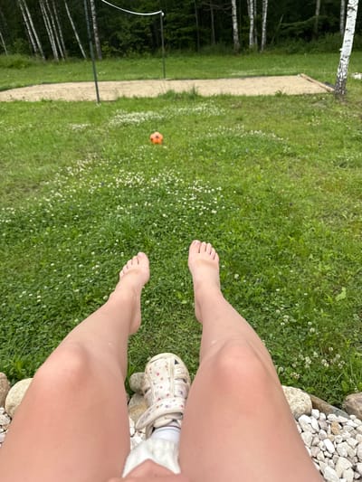 The image shows a first-person perspective of a person sitting on a rocky area, with their legs extended forward. The person is barefoot, wearing one sandal with a floral pattern on their right foot. Their left foot is visible and bare. The background consists of a grassy area with small white flowers, a sand court with a net, and a line of trees. An orange ball is visible on the grass in the distance. The environment appears to be an outdoor recreational area, possibly a backyard or park. The location is identified as Ryzhkovo, Russia.