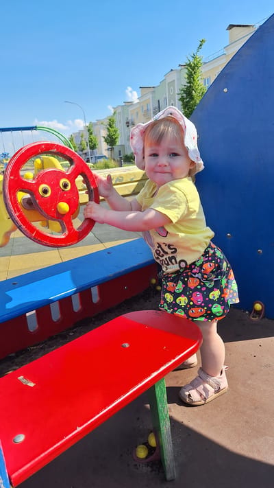 A young child with blonde hair, wearing a pink sun hat, yellow t-shirt, and a colorful skirt with cat prints, stands at a playground on a sunny day. The child is holding onto a red steering wheel, appearing to play on a toy vehicle. The playground has blue and red structures, and in the background, there are residential buildings and trees. The ground is covered with sand and wood chips. The child is facing the camera and smiling.