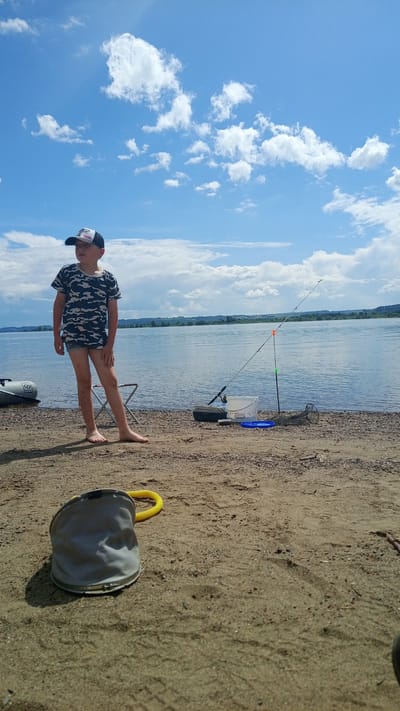 The image depicts a young boy standing barefoot on a sandy and pebbly shore beside a wide body of water in Шолья, Russia. The boy, dressed in a camouflage t-shirt, light shorts, and a backward baseball cap, is positioned slightly left of center, looking towards the water or the distant horizon under a bright, partly cloudy sky. To his right, a fishing rod is set up on a bank stick, extending over the water, accompanied by a white plastic bucket, a dark tackle box, a blue disc, and a coiled mesh fishing net. An inflatable boat is partially visible near the water's edge to the boy's left, and a small folding chair is behind him. In the immediate sandy foreground, a collapsed gray bucket with a yellow handle rests on its side. The background features a distant shoreline with faint green vegetation and low hills beneath a horizon where the sky meets the water.