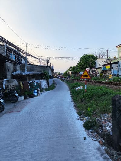 The image depicts a narrow concrete access road running parallel to a single railway track in Thành phố Quảng Ngãi, Vietnam. The road, characterized by horizontal surface grooves, gently curves towards the left into the distance. On the left side of the road, a densely built-up area features several weathered residential structures with multi-story facades, corrugated metal roofing, and exposed utility wiring. Two motorcycles are partially visible parked on the far left foreground. Assorted debris, containers, and potted plants are distributed along the base of these structures.
On the right side of the road, a grassy embankment ascends to a single set of railway tracks laid on gravel ballast. A prominent triangular railway crossing warning sign, featuring a red border, yellow background, and a black train symbol, is positioned on a red and white striped pole between the road and the tracks. Beyond the railway, additional residential buildings, interspersed with green trees and displaying various signs, line the horizon. The sky is clear, exhibiting a light blue hue indicative of daytime. No individuals, specific actions, or interactions are observable within the frame.