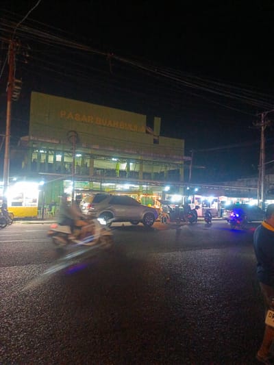 The image depicts a street scene at night in Luwuk, Indonesia. A large, two-story building with green facade and "PASAR BUAH BULILA" written in yellow is prominent in the background. The building appears to be a market. The street is paved and wet, reflecting the lights from various stalls and vehicles. Several people are visible, some on motorcycles, others standing near stalls. A silver SUV is parked on the street. There are food stalls with bright lighting set up along the side of the road. Overhead, power lines crisscross the dark sky. The overall impression is of a busy, illuminated marketplace at night.