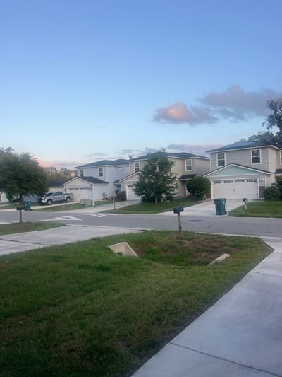 The image depicts a suburban street scene in Jacksonville, Florida, United States of America, during early morning or late evening with a clear sky and soft pastel-colored clouds. The foreground features a concrete sidewalk and a grassy area with a drainage culvert. In the middle ground, a paved road curves through a residential neighborhood lined with modern two-story houses. These houses are painted in light blue and gray tones, with white trim and garages. A silver pickup truck is parked in a driveway. Green trash bins are visible on the sides of the road. The background shows more houses and trees, with the sky taking up the upper portion of the frame. The overall impression is one of a quiet, well-maintained residential area.