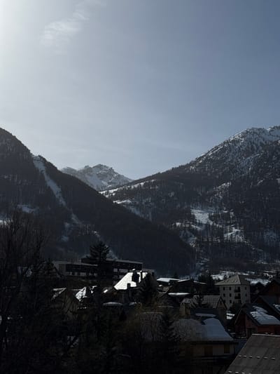 The image presents an elevated view of the city of La Salle-les-Alpes, France, under winter conditions. The upper third of the frame is occupied by a bright, hazy, light blue sky with a prominent light source in the upper-left quadrant. The background features rugged, snow-covered mountains. A steep, forested mountain with visible cleared ski runs or paths dominates the left side, while another heavily forested mountain with snow dusting rises on the right. A distinct, snow-capped rocky peak is centered in the mid-distance between the primary mountain ranges.
The mid-ground and foreground depict a section of the La Salle-les-Alpes village. Numerous structures are visible, including multi-story buildings with various roof types, many of which are covered in snow. A rectangular, multi-story building is prominent in the mid-left, partially obscured by bare deciduous trees and evergreen trees. The foreground is heavily shadowed by a dense canopy of bare trees and other vegetation, obscuring ground details. The lighting creates strong contrast, with bright sunlight illuminating the sky and upper mountain areas, while the village and lower slopes remain largely in deep shadow. No individuals or active subjects are discernible within the scene.