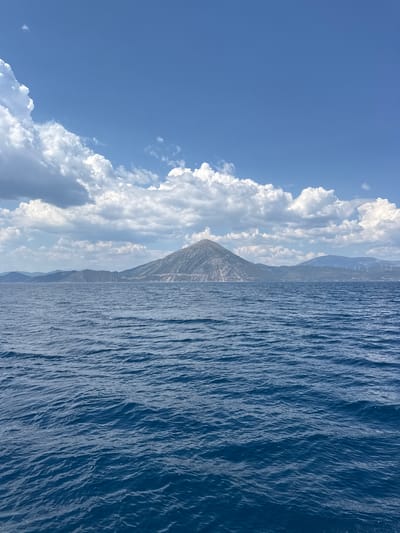 The image depicts a wide view of the sea with a mountain range in the distance. The water is a deep blue with small waves. The sky is a bright blue with scattered white clouds. A large, conical mountain dominates the center of the background, with signs of quarrying or construction on its slopes. To the right, other mountain ranges are visible, and on one of them, wind turbines can be seen. The scene appears to be a coastal landscape, likely somewhere in Greece, given the mention of Patras.