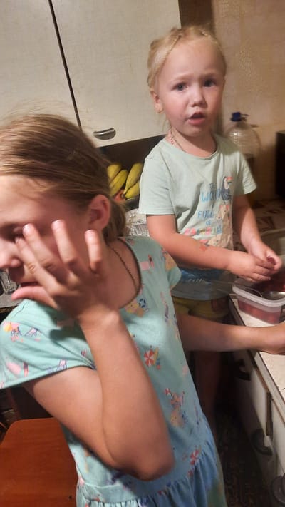 The image depicts two young girls in what appears to be a kitchen setting. The girl in the foreground, with blonde hair, is partially obscured by her hand, which is raised towards her face. She is wearing a light blue dress with a pattern of beach umbrellas and deck chairs. The girl in the background, also with blonde hair styled in braids, is facing the camera. She is wearing a light green t-shirt with a graphic print of a toucan and text that reads "The World is Full Of." She has a small earring in her left ear and a necklace. Her hands are engaged in an activity involving a container of red berries and a spoon. In the background, there are bananas, a bottle of water, and kitchen cabinets. The environment suggests a domestic interior. The image does not contain any information to indicate the location is Samar, Ukraine.