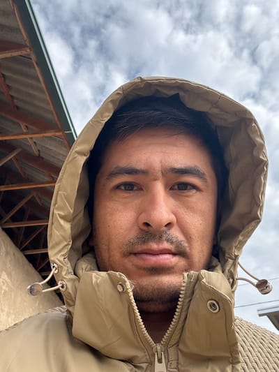 A man with dark hair and a light beard peers into the camera, wearing a beige hooded jacket. The hood is pulled up around his head. The sky is visible in the background, with a few clouds. Part of a building structure with a corrugated roof is visible in the lower left corner. The zipper of the jacket is partially open.