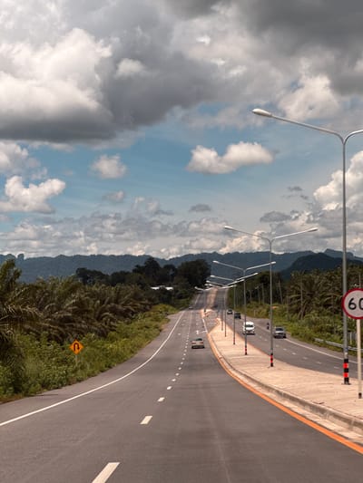 This is a photograph of a multi-lane highway in Plai Phraya, Thailand. The highway curves to the left and ascends into the distance. On the left side of the road, there are palm trees and lush green vegetation, with a yellow U-turn sign visible. On the right side of the road, there is a paved median with a concrete barrier and lampposts lining the highway. A speed limit sign indicating "60" is posted on the right. Several cars and a motorcycle are present on the highway. In the background, a range of mountains covered in dense forest can be seen under a sky with scattered clouds.