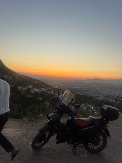 The image, taken at sunset, captures a scenic view from a high vantage point overlooking Tétouan, Morocco. The sky displays a striking gradient, with a vibrant band of orange and yellow hues along the horizon, transitioning upwards into a softer blue and grey. Distant mountains are silhouetted against the colorful sky, behind which a sprawling city or townscape is visible, with numerous buildings and early evening lights beginning to illuminate the urban expanse.

In the foreground, a dark-colored motorcycle with subtle red detailing on its seat is prominently parked on a dirt or gravel path to the right. It appears to be equipped with a black top case or luggage on its rear rack, suggesting it may have been used for travel or a ride to this elevated spot. A partial view of a person is seen in the bottom left corner, showing their lower torso, legs, and feet, indicating they are standing near the motorcycle, facing the panoramic view.

The scene conveys a sense of peace and contemplation at the close of day, with the clear sky suggesting fair weather. There is no visible text in the image.
