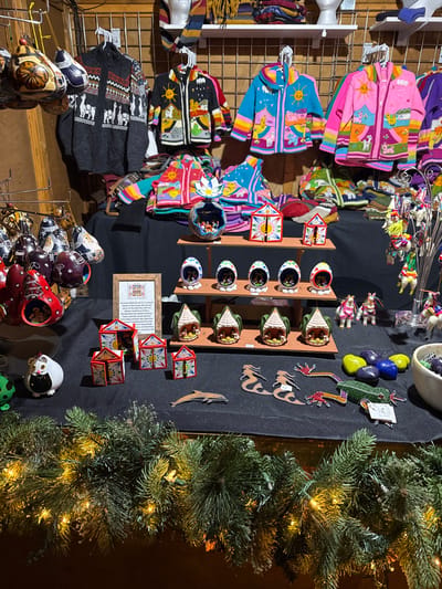 A table display features an array of colorful handcrafted items. Clothing, including patterned sweaters and jackets, hangs on a wire rack. Ornaments and painted gourds are arranged on the table, along with wooden animal figures. A strand of green garland with small lights adorns the right side of the table.