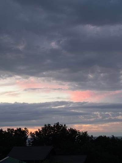 The image depicts a sky with dramatic cloud formations at dusk, likely over a natural landscape. The clouds are predominantly dark gray, with streaks of pale pink and orange suggesting the last rays of sunlight breaking through. The overall atmosphere is moody and serene.

In the foreground and to the right, silhouetted against the sky, are dense trees with dark foliage. A dark-tiled roof of a building is partially visible on the far right, hinting at a semi-rural or suburban setting. The horizon line is low, with the sky occupying the majority of the frame. The time of day is clearly sunset or twilight due to the colors in the sky and the silhouettes. The weather appears to be clear or partly cloudy, with the clouds contributing to the visual drama rather than indicating imminent rain.

No people or specific objects are clearly discernible besides the natural elements and the hint of a structure. The image evokes a sense of calm and the quiet beauty of nature transitioning from day to night. The location is identified as Voronov, Russia. No text is visible in the image.