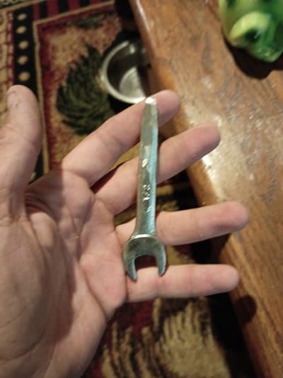 A silver-colored wrench is being held in a right hand. The hand has fair skin. The background includes a wooden surface, a decorative rug with floral and geometric patterns, and a small, green, potentially glass, object. The location is not specified in the image. There is no information suggesting the city of Clarksville, United States.