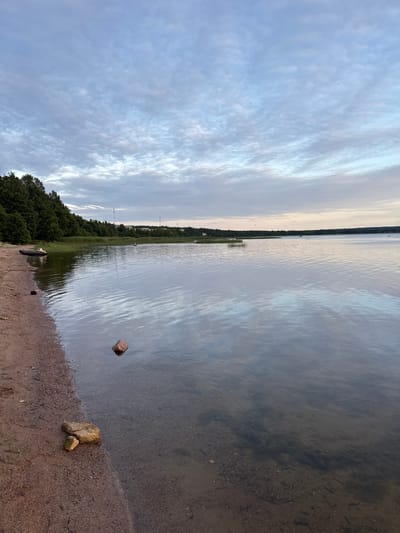 The image depicts a serene lake scene on the outskirts of Glebychevo, Russia. The water is calm, reflecting the overcast sky and the dense green treeline that borders the opposite shore. In the foreground, a sandy and pebbly beach is visible, with a few scattered rocks. An inflatable boat, dark gray in color, rests on the shore. The sky is filled with a layer of scattered, pale clouds. The overall impression is one of a tranquil, natural landscape.
