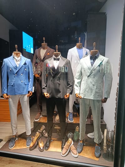 The image shows a display window of a men's clothing store. Five mannequins are dressed in suits and accessories. From left to right, the first mannequin wears a blue pinstriped double-breasted suit with a white shirt and a polka dot ascot. The second mannequin wears a brown suit with an orange pocket square and a beige shirt. The third mannequin is dressed in a black tuxedo with a bow tie and a white shirt. The fourth mannequin wears a light blue collared shirt and a light blue blazer. The fifth mannequin is dressed in a light grey linen double-breasted suit with a dark blue shirt and a green pocket square.
Various men's shoes are displayed on wooden platforms in front of the mannequins, including loafers, oxfords, and sneakers in shades of brown, black, blue, and green. Belts are also displayed on a stand to the right. The background appears to be the interior of the store, with shelving and clothing racks visible. There is a large screen displaying an image on the left side of the window.
The reflection on the glass suggests the photo was taken from across the street. The store is located in Niš, Serbia, specifically on Obrenovićeva street, a well-known pedestrian zone in the city center, home to numerous retail establishments. The overall impression is a well-organized and stylish display of men's formal and casual wear.