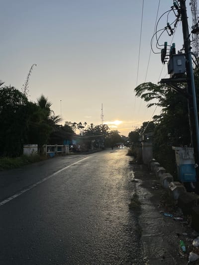 This media file captures a street scene in Cotseutui, Indonesia, likely during a late afternoon or early evening. The perspective is from the side of a wet asphalt road, reflecting the golden light of a setting sun. Overhead, a complex network of electrical wires and utility poles dominates the left side of the frame, with a transformer box visible. Dense foliage and palm trees line the road, interspersed with small buildings and structures. A communication tower is visible in the distance. There are a few vehicles on the road, suggesting some activity, but no people are clearly discernible. The presence of scattered litter along the roadside, including plastic bottles and wrappers, indicates a lack of regular maintenance in that area. The overall mood is somewhat subdued, with the setting sun casting long shadows and the wet road adding a sense of dampness. No visible text is present in the image.