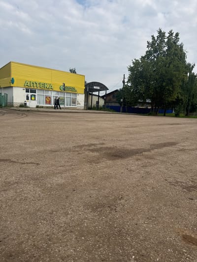 In Chastyye, Russia, a low-angle shot captures the exterior of a bright yellow building with "АПТЕКА" (Pharmacy) and "ПЛАНЕТА ЗДОРОВЬЯ" (Planet of Health) signage in large green letters. The building has a white base with stone accents and large windows, some displaying signs with percentages and medical-related imagery. Two individuals, a man and a woman, are walking away from the building on a sidewalk in front of it. To the right of the pharmacy, a covered structure with a white roof and support beams is visible. Beyond the building, mature trees with dense green foliage are prominent. The foreground consists of a wide, uneven, light-brown paved area, possibly a parking lot or open space, with visible tire marks and darker patches indicating moisture or wear. The sky above is overcast with scattered white clouds. A street lamp and a dark blue fence can be seen in the middle ground behind the pharmacy and trees. The overall scene depicts a commercial area in a town.