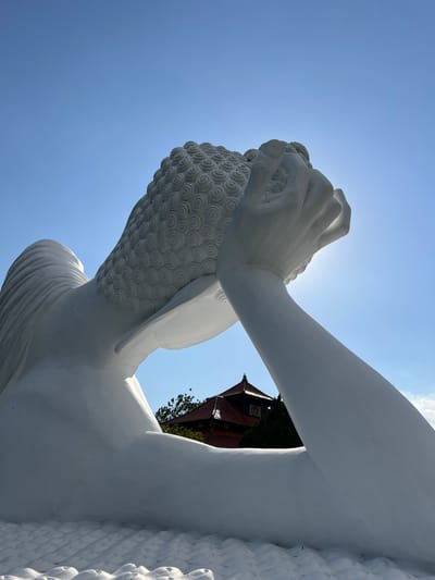 A low-angle, close-up, outdoor, eye-level shot showcases a large, white statue of a reclining Buddha against a bright blue sky with a hint of the sun creating a slight glare. The sculpture is white and appears to be made of smooth, polished material. The Buddha's head is tilted back, resting on one hand which is positioned under its head. Its hair is rendered in intricate, swirling patterns. The body of the Buddha is partially visible, with a flowing robe draped over its shoulder. In the background, a traditional building with a red tiled roof is visible amongst green trees, and a glimpse of a circular clock face can be seen on the building. The foreground shows the textured, wave-like pattern of the base of the statue. The overall impression is one of peaceful repose and grandeur. The image was taken in Pupuan, Indonesia.