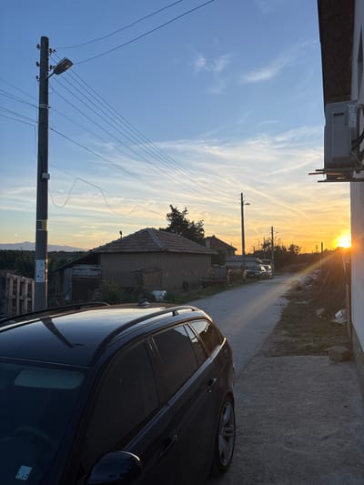 The image depicts a rural street scene in Rosen, Bulgaria, at sunset. A dark-colored station wagon is parked in the foreground on the left, its roof and side visible. The street is paved and curves to the right, leading towards the setting sun. Two other cars are parked further down the street. On the left side of the street, a utility pole with a street light and numerous wires stands tall. Beyond the pole, several traditional houses with tiled roofs are visible, surrounded by vegetation. The right side of the street features a white-walled building with an air conditioning unit mounted on its exterior. Debris and construction materials are scattered along the side of the road. The sky is a gradient of blue and orange, with wispy clouds suggesting the sun is low on the horizon. In the distance, mountains are faintly visible.