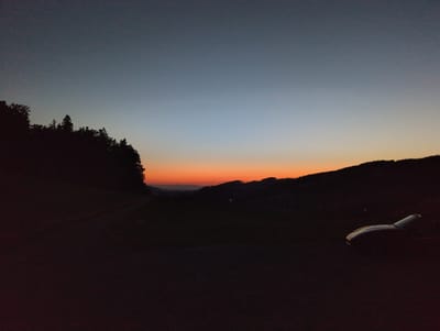 The image depicts a twilight scene, likely in the municipality of St. Peter, Germany, characterized by a gradient of colors in the sky transitioning from a deep blue at the top to warm orange and red hues near the horizon. Silhouetted against this colorful sky are rolling hills and a dense line of trees on the left side of the frame. In the foreground, a dark, unpaved path winds through the landscape. On the right side of the image, the silhouette of a car is visible, parked or stopped near the edge of the path. The overall impression is one of a serene, natural environment at dusk.