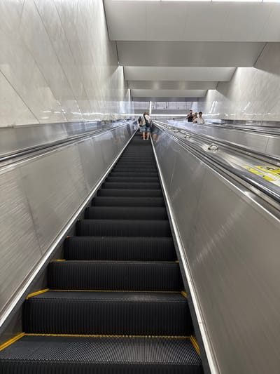 The image shows a downward-facing view of a modern, well-lit escalator in what appears to be a subway station or a large public building. The escalators have black treads with yellow safety strips and are flanked by brushed metal sides. Several people are visible on the escalators, some ascending and some descending, with their backs mostly to the camera. The walls of the structure are clad in large, light-colored tiles, and the ceiling is a series of white geometric panels. The overall impression is one of a clean, contemporary public space. Without further information, the specific location in Baiyun District, China cannot be determined from the visual evidence alone.