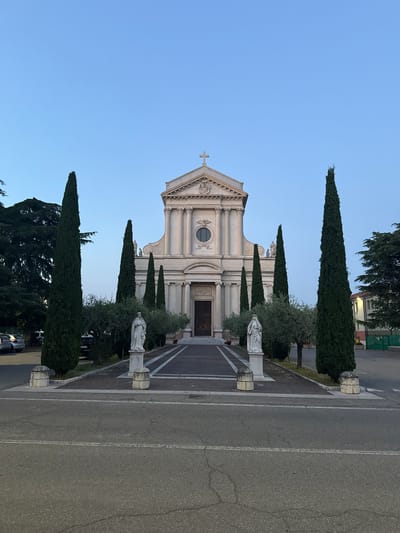 The image shows the Church of Saints Peter and Paul in Villafranca di Verona, Italy. The facade of the church is light-colored with a triangular pediment, a central circular window, and a cross atop the pediment. On either side of the pathway leading to the church entrance are statues on pedestals, positioned amidst trees. Tall, slender cypress trees line the path, adding verticality to the scene. The foreground is dominated by a paved road with lane markings, and the sky is a clear blue.