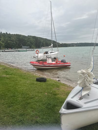 An overcast day at a lake in Chmielno, Poland, features a shoreline with grass and a sandy edge. Moored boats are visible on the water, including a white sailboat with a mast, a red and white inflatable rescue boat with "ASEKUKANT" and "KOLBUDY" written on it, and a portion of a small gray sailboat in the foreground. The rescue boat has an outboard motor and a life preserver attached to its side. A deflated black tire rests on the grass near the water's edge. In the background, a tree-lined shore with some structures on a dock is visible.