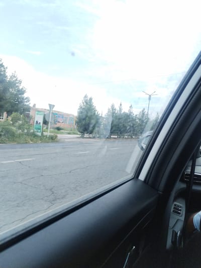 The image is taken from inside a vehicle, looking out the passenger window. The view shows a street with a cracked asphalt road. On the left side of the road, there is a sidewalk with some greenery and trees. Beyond the sidewalk, a building with an orange facade is visible, adorned with colorful murals. A traffic sign is also present near the building. In the background, more trees and a street light are discernible against a bright, cloudy sky. The interior of the car is visible in the foreground, showing the door panel and a person's leg wearing light-colored shorts. The setting appears to be the city of Istaravshan, Tajikistan.