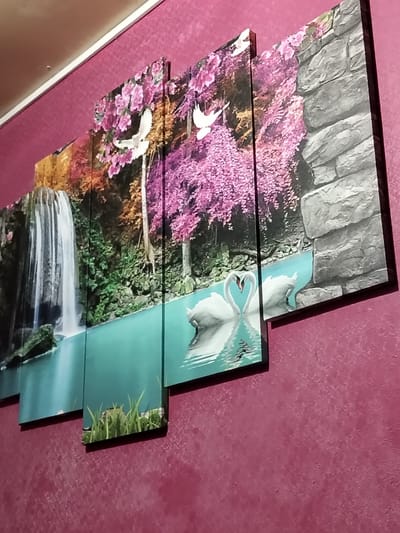 This is a canvas print of a serene natural scene, displayed on a textured pink wall. The artwork is a multi-panel piece, divided into five sections that together create a panoramic view. The scene depicts a waterfall cascading down mossy rocks on the left, transitioning into a vibrant forest with trees adorned with abundant pink and orange foliage. Two white doves are gracefully flying amidst the colorful blossoms, adding a touch of ethereal beauty. In the foreground, a tranquil turquoise body of water reflects the surrounding scenery. Two white swans are swimming in the water, their necks curved to form a heart shape, symbolizing love and peace. The rightmost panel features a textured stone wall, adding an element of rustic charm to the overall composition. The lighting suggests a bright, perhaps late afternoon, setting, with a sense of calm and natural splendor. There is no visible text in the image. The scene is likely meant to evoke feelings of tranquility and romance.