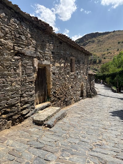 This is a street scene in Patones De Arriba, Spain, a historic village known for its slate architecture. The image showcases a cobblestone street winding uphill alongside a weathered stone building. The building's facade is constructed from rough-hewn, irregular slate stones, with a wooden door and a small, arched window visible. The roof appears to be made of terracotta tiles.
The street itself is paved with dark grey cobblestones, showing signs of age and wear. To the right of the building, a green vine-covered pergola creates a shaded walkway. Further up the street, two figures, a man and a woman, are walking away from the camera, casting shadows on the cobblestones. In the background, a dry, hilly landscape under a bright blue sky with scattered white clouds completes the scene. A vintage-style streetlamp is mounted on the stone wall. The overall impression is one of rustic charm and timeless beauty, characteristic of Patones De Arriba.