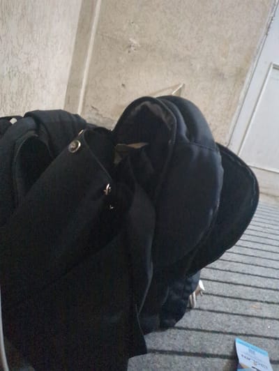 The image shows a black backpack on a textured surface. The backpack is partially open, revealing some of its contents. In the background, there is a striped surface and a sliver of a white object with text, possibly a ticket or card. The scene appears to be outdoors, with a light-colored wall in the upper left corner. The lighting suggests it could be daytime. The overall impression is of a casual, everyday scene.