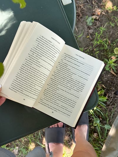 An open book is placed on a dark green surface, likely a table, outdoors. The book, titled "Ріо Штовхає Мене Плечем" on the right page, lies open to pages 94 and 95. The pages are filled with Ukrainian text. A person's hand is visible on the left side of the book, holding it open. In the background, out of focus, is a grassy area with some plants and the hint of a tree, suggesting a natural or park-like setting. The ground appears to be soil. The image is taken from a slightly elevated perspective, looking down at the book and the surrounding environment. The scene is illuminated by natural light, indicating it is daytime. The location is specified as Yablunivka, Ukraine.