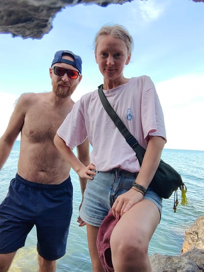 The media file features a medium shot of a man and a woman posing for a selfie in what appears to be a coastal setting. The man, on the left, is shirtless, wearing dark blue swim trunks and a blue baseball cap. He has a beard and is wearing sunglasses. The woman, on the right, is wearing a light pink t-shirt and denim shorts. She has a dark green fanny pack slung across her body and is holding a maroon hat. The background shows a bright blue sky with some white clouds and the calm, clear blue sea. The overall atmosphere suggests a warm, sunny day, possibly during the daytime. The couple appears relaxed and content, looking towards the camera with smiles. The foreground is partially obscured by a rocky edge and the water, indicating they are standing near the shore. The location context provided is Utios, Ukraine. There is no visible text in the image.