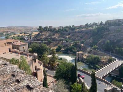 This is an eye-level, outdoor, wide shot of Toledo, Spain, taken on a clear, sunny day with a few scattered clouds. The foreground is dominated by a textured, weathered stone wall and some overhanging foliage.
In the mid-ground, several buildings with terracotta-colored tile roofs are visible, some with multiple stories and balconies. A winding river flows through the scene, with a small waterfall creating white foam. On the riverbank, a historic, weathered stone building with multiple windows and a pitched roof stands. A modern red SUV is driving on a paved road that curves along the riverbank and leads to a bridge.
The background features a steep, rocky hillside covered in dry vegetation and scattered trees. Several ancient-looking stone towers are situated on top of the hill, along with a long stone wall and more trees. The landscape stretches out into a dry, rolling terrain with distant buildings. A modern bridge with multiple lanes crosses the river in the background, connecting to a road that winds up the hillside. Streetlights are visible along the roads. The overall scene depicts a historic Spanish city with a river, ancient architecture, and modern infrastructure.