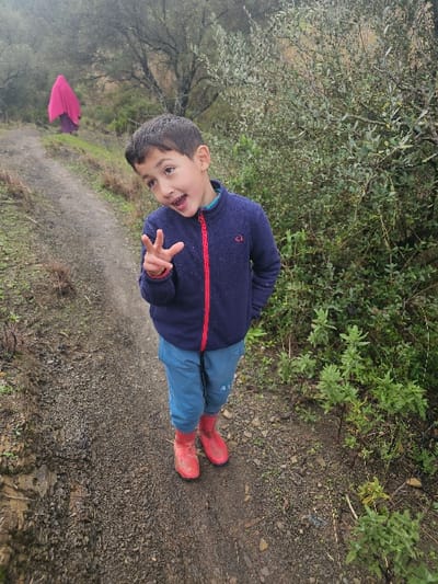 The image captures an outdoor scene on a dirt path in a natural, vegetated area near Moqrisset, Morocco, under overcast conditions. In the foreground, a young boy, approximately 5-8 years old, with dark hair, stands facing the camera. He wears a dark blue fleece jacket with a red zipper, light blue pants, and bright red rubber boots. His mouth is open in a wide smile, and he holds his right hand up in a "peace" sign, while his left hand is in his pocket. Behind the boy, further up the winding dirt path, an adult figure is seen from behind, walking away from the camera. This individual is enveloped in a bright fuchsia/magenta garment, covering their head and body. The path itself is unpaved, muddy, and strewn with small rocks, flanked on both sides by dense green shrubs and trees that extend into the background.