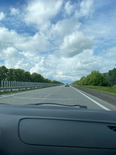 The image depicts a view from inside a vehicle traveling on a highway, likely in or near Subai, Russia, given the context. The road is paved and divided into lanes by white markings. A white car is visible in the distance, traveling in the same direction. On the left side of the road, there is a guardrail composed of vertical concrete posts with reflective red markers and a horizontal metal beam. Beyond the guardrail, a dense line of green trees forms a natural barrier. The right side of the road also features a grassy verge and more trees. The sky above is a mix of blue sky and large, fluffy white clouds. The foreground shows the dark interior of the vehicle's dashboard and windshield wipers.