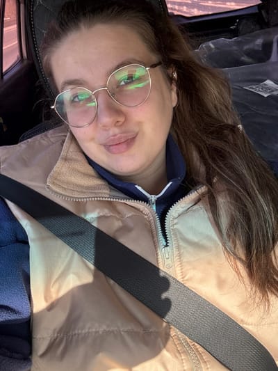 A young adult female is depicted seated in the passenger side of a vehicle in the city of Kukushtan, Russia. She has light skin, long brown hair, and is wearing round, metallic-rimmed glasses. Her facial expression shows a slight smile with pursed lips, and she looks directly towards the camera. She wears a light beige or tan puffer vest over a dark blue long-sleeved top with a light blue zipper. A dark grey seatbelt crosses her chest diagonally from her left shoulder to her right hip. The vehicle's interior is visible, including a dark headrest behind her and an out-of-focus window showing a blurred exterior scene to her left. Natural light illuminates her from the left side, creating a shadow across her chest from the seatbelt.
